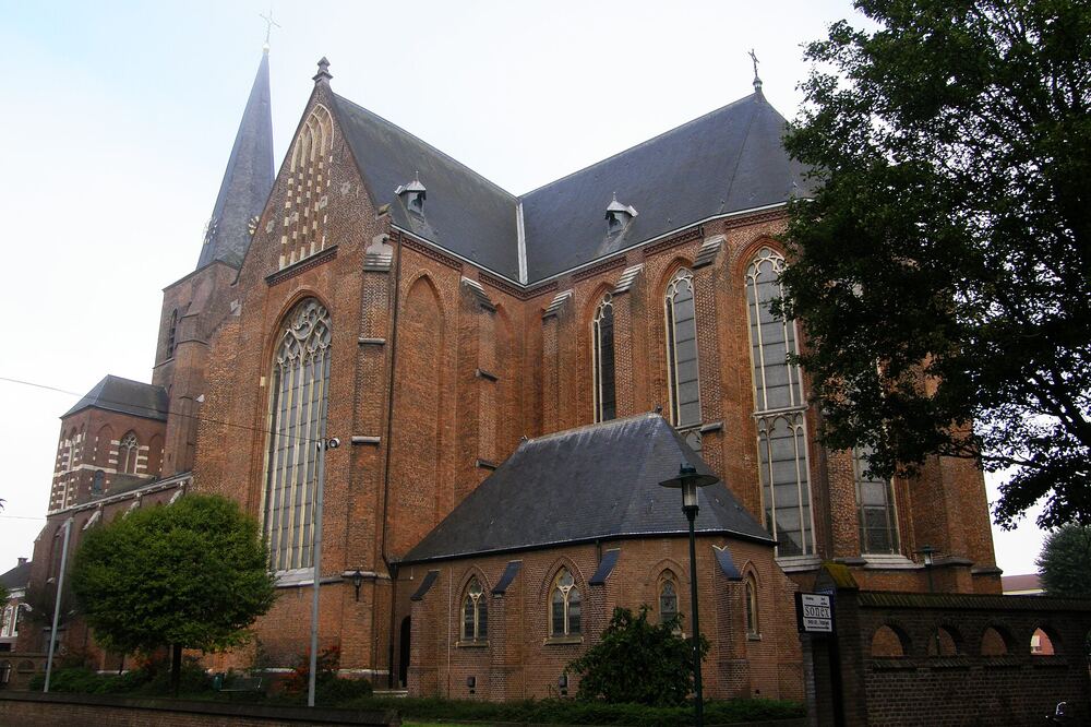 De bakstenen Sint-Willibrorduskerk in Deurne, met spitse toren, grote gotische ramen en een lage kerkhofmuur ervoor